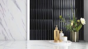Sleek black tiled wall with vertical ridges behind elegant gold and white bathroom accessories, including a soap dispenser and a vase with fresh flowers, creating a luxurious modern bathroom decor.