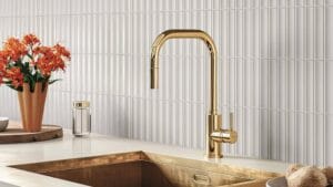 Luxurious gold kitchen faucet with modern curved design, installed above a copper-colored sink, surrounded by white tile backsplash, decorative flowers, soap dispenser, and natural stone in a stylish kitchen setting.