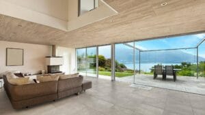 Spacious modern living room with large glass sliding doors showcasing a lake and mountain view, featuring neutral-colored furniture, a fireplace, and a wooden ceiling.