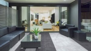 Modern outdoor patio with black leather seating, glass coffee table, and decorative plant, leading into a stylish open-concept living room with wooden flooring and contemporary furniture, ideal for outdoor and indoor living spaces.