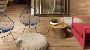 Colorful outdoor patio furniture with modern chairs, a woven ottoman, and a side table featuring potted plants and drinks, set against a wooden deck and glass door backdrop.