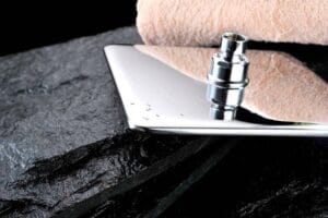 Shiny modern kitchen sink faucet with water droplets on a sleek chrome surface, placed on a black stone countertop, with a pink towel in the background. Ideal for bathroom or kitchen renovation projects.