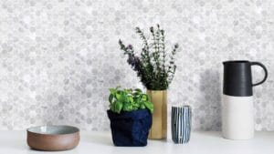 Vase with purple flowers and green foliage, black and white ceramic pitcher, striped ceramic cup, and brown ceramic bowl on a white surface against a hexagonal tile background.