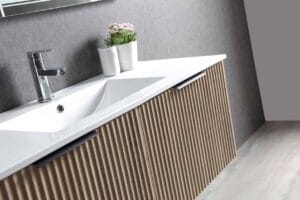 Contemporary bathroom vanity with wooden slat cabinet, white countertop, built-in sink, modern chrome faucet, and decorative potted flowers against a gray textured wall.