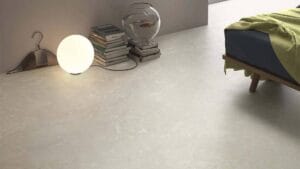 Polished beige stone floor tiles in a modern, minimalistic bedroom corner. Features include a small wooden bed with green and navy bedding, a glowing round table lamp, a stack of books, and a decorative fishbowl.