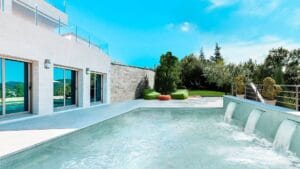 Modern outdoor swimming pool with waterfall feature, adjacent to a contemporary house with large glass doors, surrounded by lush greenery and outdoor seating for relaxation.