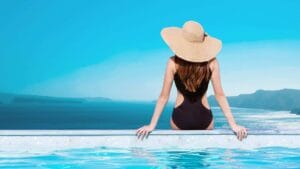 Relaxing woman in a black swimsuit sitting by the poolside with a wide-brimmed sun hat, overlooking a scenic ocean view on a bright sunny day.